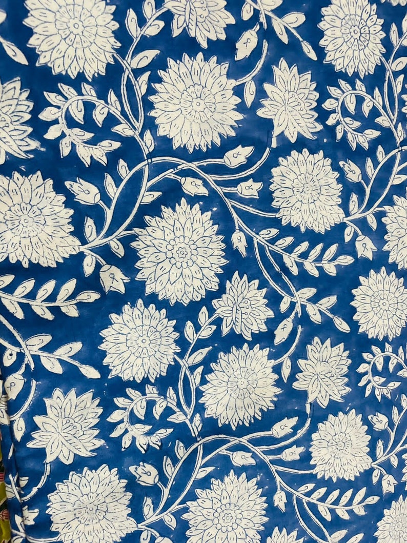 May include: A blue fabric with a white floral pattern. The flowers are stylized and have a circular shape with multiple layers of petals. The fabric has a vintage look and feel.