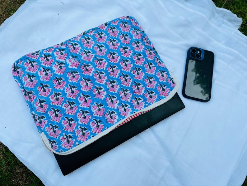 May include: A blue and pink floral laptop sleeve with a white trim. The sleeve is folded over a black laptop and a black phone is lying on a white surface.