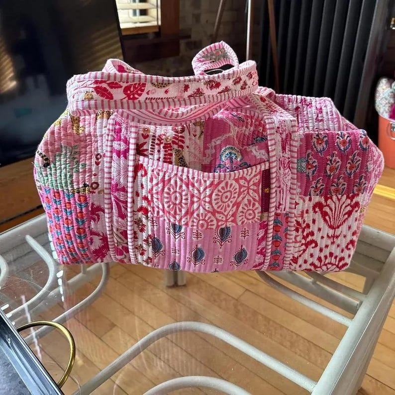 May include: A patchwork tote bag in shades of pink, red, and white. The bag features various floral and geometric patterns, with a quilted texture. It has multiple pockets and a top handle.