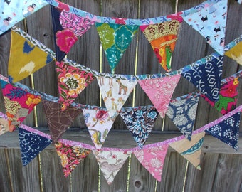 May include: A colorful fabric banner with triangular flags featuring various patterns and designs. The flags are sewn together with a pink ribbon. The banner is hanging on a wooden fence.