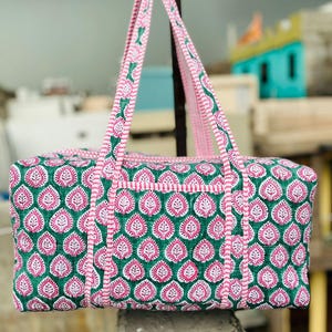 May include: A quilted tote bag with a green and pink floral pattern. The bag has two pink handles and a matching trim. The bag is rectangular with a top zipper and a small pocket on the front.