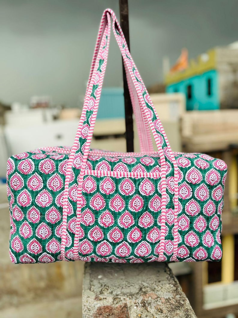 May include: A quilted tote bag with a green and pink floral pattern. The bag has two pink handles and a matching trim. The bag is rectangular with a top zipper and a small pocket on the front.