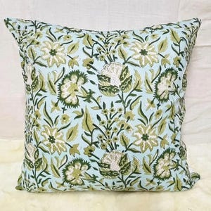 K&ouml;nnte beinhalten: Ein quadratisches Dekokissen mit hellblauem Hintergrund und einem sich wiederholenden Muster aus dunkelgr&uuml;nen und olivgr&uuml;nen Blumenmotiven, einschlie&szlig;lich stilisierter Bl&uuml;ten, Bl&auml;tter und St&auml;ngel in einem symmetrischen botanischen Design.