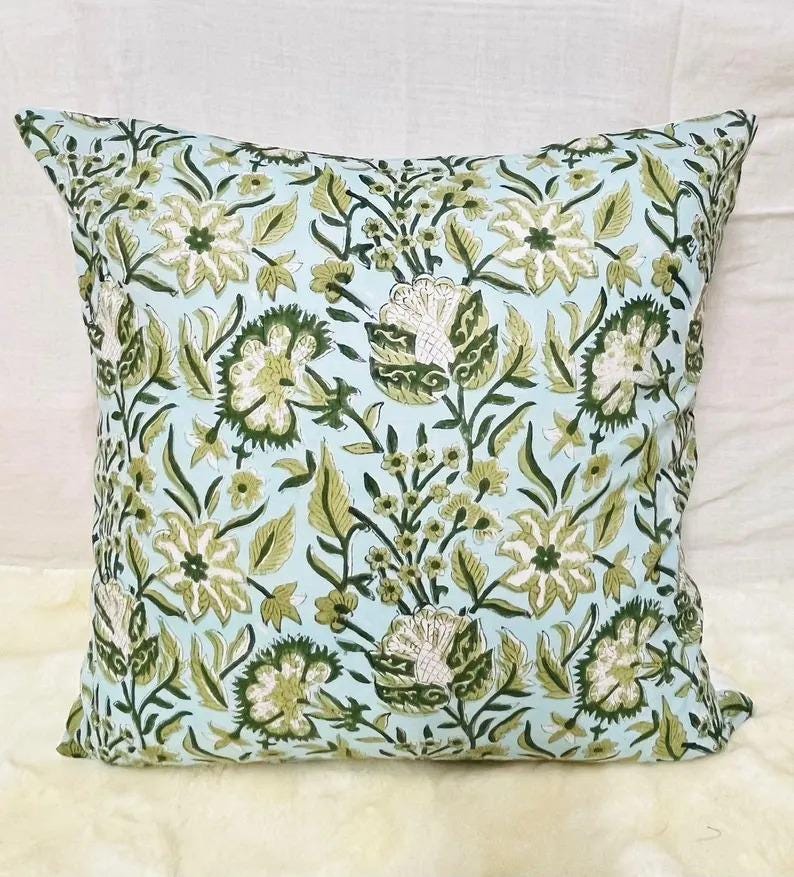 K&ouml;nnte beinhalten: Ein quadratisches Dekokissen mit hellblauem Hintergrund und einem sich wiederholenden Muster aus dunkelgr&uuml;nen und olivgr&uuml;nen Blumenmotiven, einschlie&szlig;lich stilisierter Bl&uuml;ten, Bl&auml;tter und St&auml;ngel in einem symmetrischen botanischen Design.