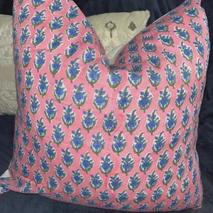K&ouml;nnte beinhalten: Ein quadratisches Zierkissen mit einem sich wiederholenden Blumenmuster aus blauen und gr&uuml;nen botanischen Motiven auf einem hellrosa Stoffhintergrund. Das Design besteht aus kleinen, stilisierten Bl&uuml;tenknospen in einem gleichm&auml;&szlig;igen Raster.