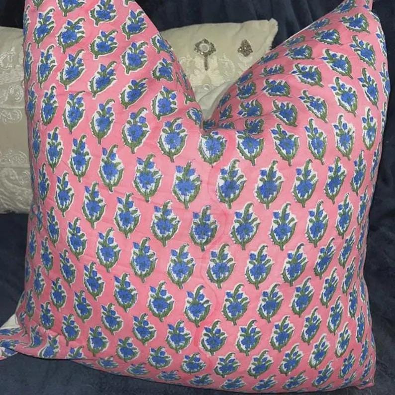K&ouml;nnte beinhalten: Ein quadratisches Zierkissen mit einem sich wiederholenden Blumenmuster aus blauen und gr&uuml;nen botanischen Motiven auf einem hellrosa Stoffhintergrund. Das Design besteht aus kleinen, stilisierten Bl&uuml;tenknospen in einem gleichm&auml;&szlig;igen Raster.