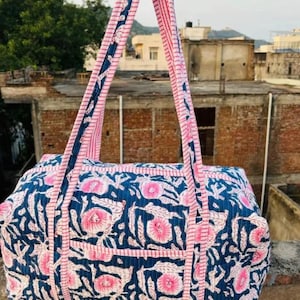 May include: A blue and pink floral print tote bag with matching handles. The bag features a rectangular shape with a pocket on the front. The handles are pink with a blue and white patterned trim. The bag is made of a quilted fabric.
