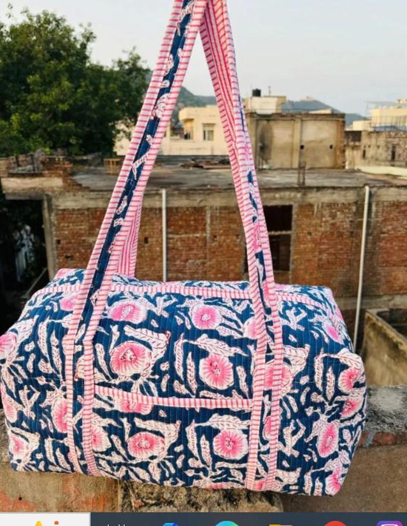 May include: A blue and pink floral print tote bag with matching handles. The bag features a rectangular shape with a pocket on the front. The handles are pink with a blue and white patterned trim. The bag is made of a quilted fabric.