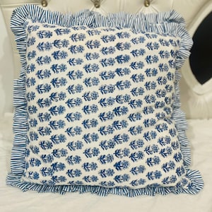 May include: A square throw pillow featuring a white base with a repeating blue floral block print pattern. The pillow is bordered by a ruffled edge made of blue and white striped fabric, creating a distinct contrast against the floral center.