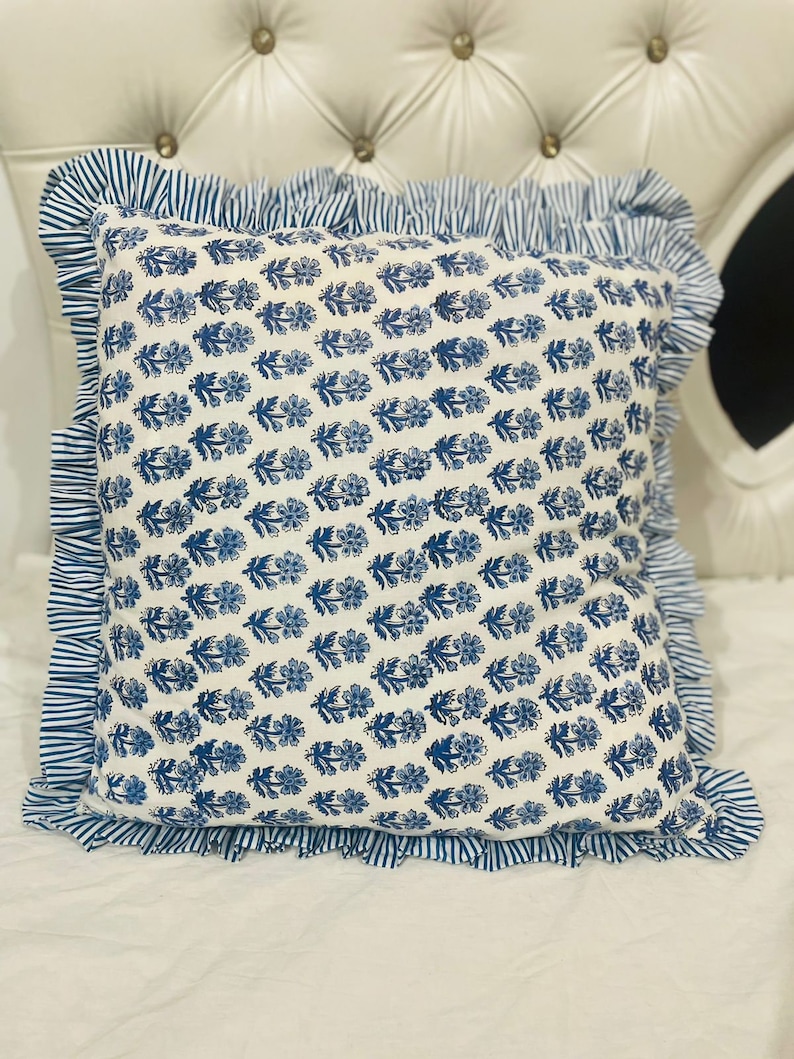 May include: A square throw pillow featuring a white base with a repeating blue floral block print pattern. The pillow is bordered by a ruffled edge made of blue and white striped fabric, creating a distinct contrast against the floral center.