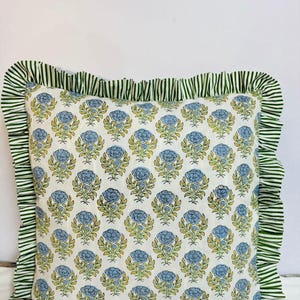 May include: A square throw pillow featuring a repeating pattern of blue flowers with green stems and leaves on a white background. The pillow is bordered by a ruffled edge made of fabric with a green and white vertical stripe pattern.