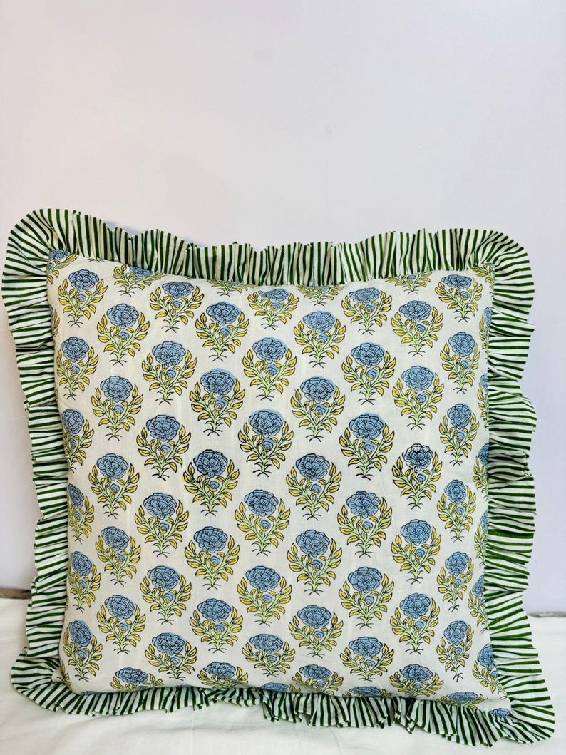 May include: A square throw pillow featuring a repeating pattern of blue flowers with green stems and leaves on a white background. The pillow is bordered by a ruffled edge made of fabric with a green and white vertical stripe pattern.