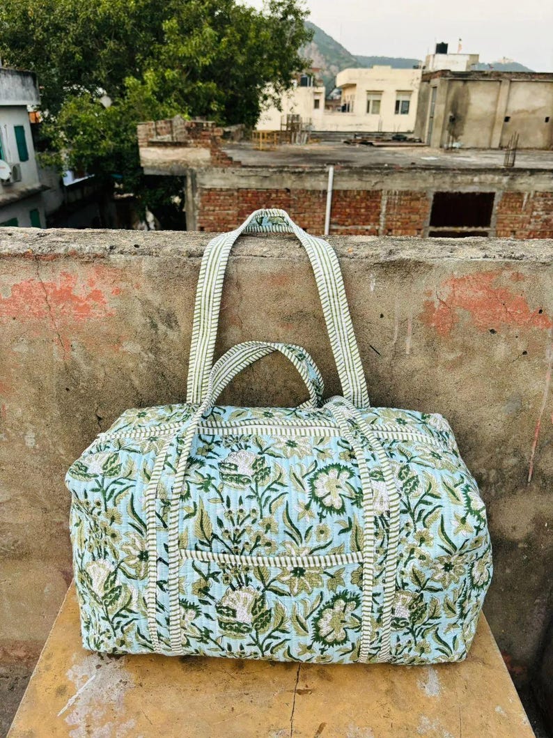 May include: A large, rectangular duffel bag with a floral pattern in shades of blue, green, and white. The bag has two handles and a striped strap. The bag is made of a quilted fabric.