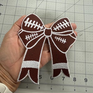 May include: A brown and white football-themed bow. The bow is made of a textured brown material with white stitching details, resembling a football. The bow is held in a hand.