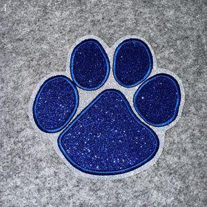 May include: A blue glitter paw print with a silver outline. The paw print is a symbol of pets and animals. The image is a close-up shot of the paw print.