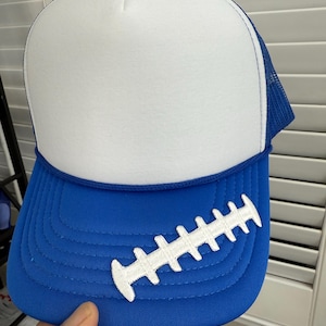 May include: A blue and white trucker hat with a football design. The hat features a white foam front panel, a blue brim, and a blue mesh back. A white football graphic is embroidered on the brim.