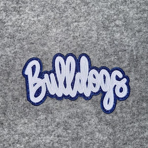 May include: A white and blue embroidered patch with the word "Bulldogs" in a cursive font. The patch has a blue border with glitter accents and is set against a gray background.