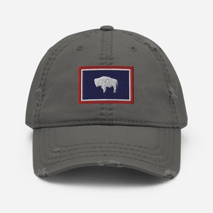 May include: A gray distressed baseball cap with an embroidered patch of the state flag of Wyoming. The flag features a white silhouette of a buffalo on a blue background with a red border.