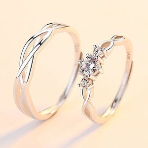 May include: Two silver rings. One ring features a woven design, while the other has a central clear gemstone flanked by smaller stones. The rings are set against a soft, neutral background, highlighting their elegant design.