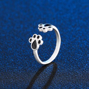 May include: A silver ring with two black paw print designs. The ring is open and adjustable.