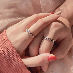 May include: Two silver rings are displayed on a person's fingers. One ring features a floral design with three clear stones. The other ring has a twisted band. The background is a soft, muted pink.