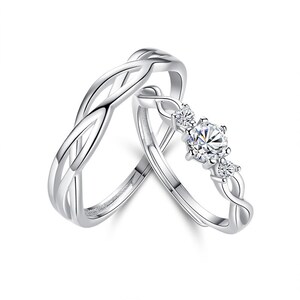 May include: Two silver rings. One ring features a braided design, while the other has a central clear gemstone flanked by smaller stones. The rings are set against a white background, highlighting their reflective surfaces.