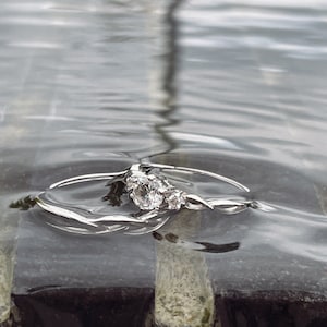 May include: A silver engagement ring with three clear gemstones, partially submerged in water. The ring has a delicate, twisted band design. The water's surface reflects the light, creating a shimmering effect. The ring rests on a dark, textured surface.