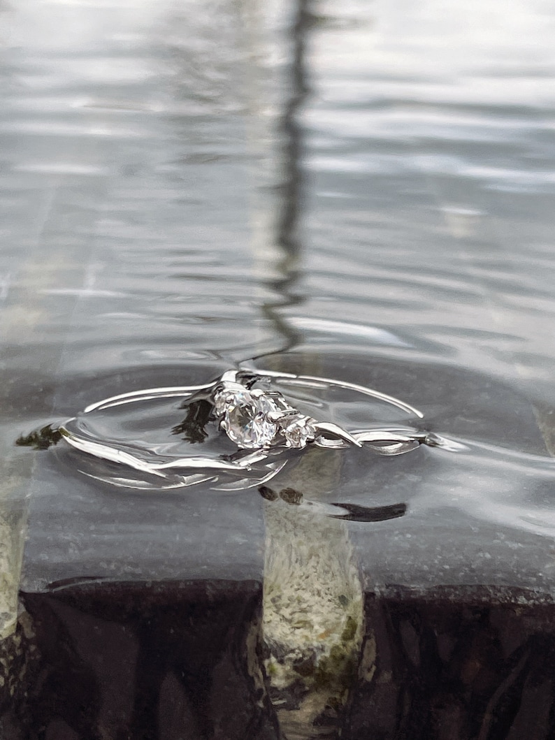 May include: A silver engagement ring with three clear gemstones, partially submerged in water. The ring has a delicate, twisted band design. The water's surface reflects the light, creating a shimmering effect. The ring rests on a dark, textured surface.