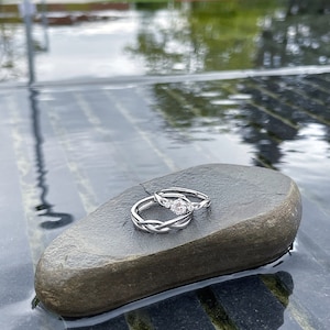 May include: Two silver rings with a braided design, one with a central gemstone, rest on a dark gray stone. The rings are set against a blurred background of water and greenery, creating a serene and elegant composition.