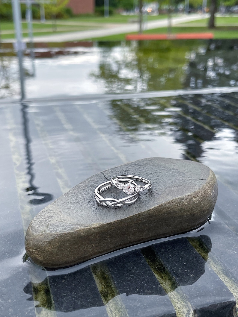 May include: Two silver rings with a braided design, one with a central gemstone, rest on a dark gray stone. The rings are set against a blurred background of water and greenery, creating a serene and elegant composition.