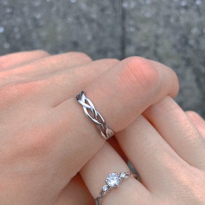 May include: Two silver rings are displayed, one with a braided design and the other with a central gemstone flanked by smaller stones. The rings are on a person's fingers, showcasing the jewelry's intricate details and design.