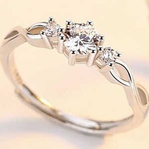 May include: A silver ring with a large, round, clear gemstone in the center, flanked by two smaller gemstones. The band features an interwoven design. The ring is set against a soft, neutral background, highlighting its sparkle and elegance.