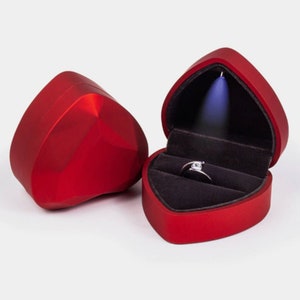 May include: Red heart-shaped ring box with a velvet interior and a ring inside. The box has a light that illuminates the ring.