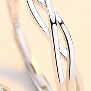 May include: A silver-colored ring with an interwoven design. The ring features a continuous, flowing pattern of intertwined bands, creating an open, airy aesthetic. The metal has a polished, reflective surface.