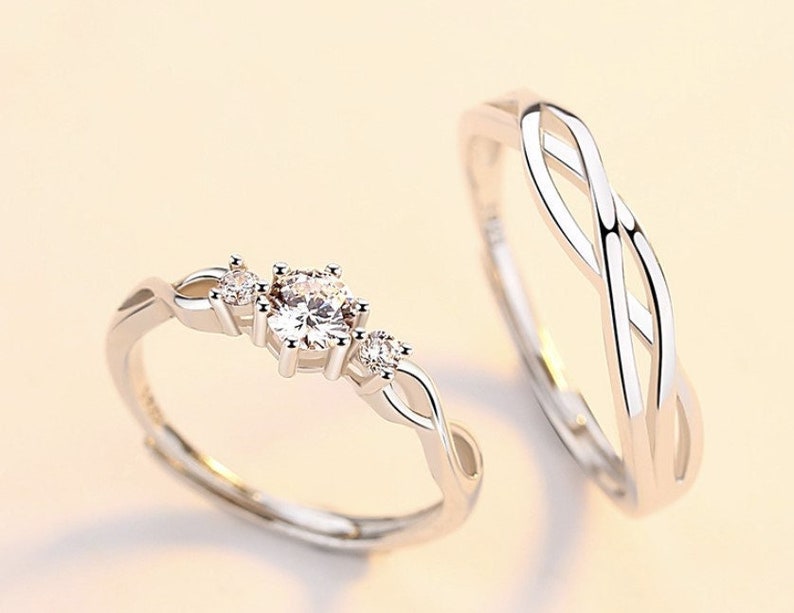 May include: Two silver rings. One ring features a central, round, clear gemstone flanked by smaller stones, set on a twisted band. The second ring has a continuous, interwoven design. Both rings are set against a light background.