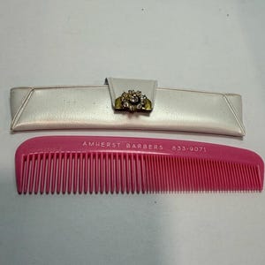 May include: A pink comb with the text "AMHERST BARBERS 833-9071" and a white case with a decorative clasp. The clasp features a gold-colored metal base with a cluster of clear and gold-colored stones.
