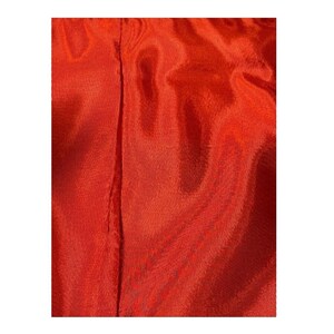 May include: Close-up of a vibrant, solid-colored fabric in a rich shade of red-orange. The material has a subtle sheen, with visible vertical seams running down the center. The texture appears smooth and flowing, suggesting a lightweight textile.