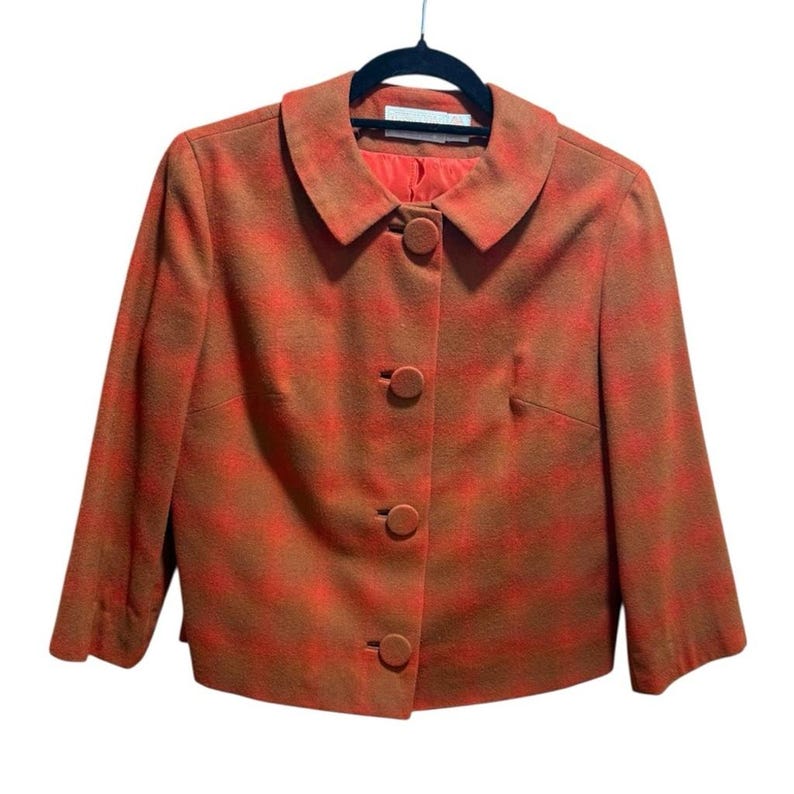 May include: A vintage orange and brown plaid blazer with a round collar and three matching buttons. The blazer is hanging on a black hanger. The interior lining is a bright orange color.