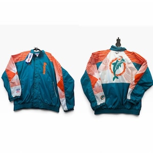 May include: A teal, orange, and white windbreaker jacket featuring the Miami Dolphins logo on the back. The front displays the team name vertically in orange, while the sleeves have orange and white striped patterns and an NFL logo patch.