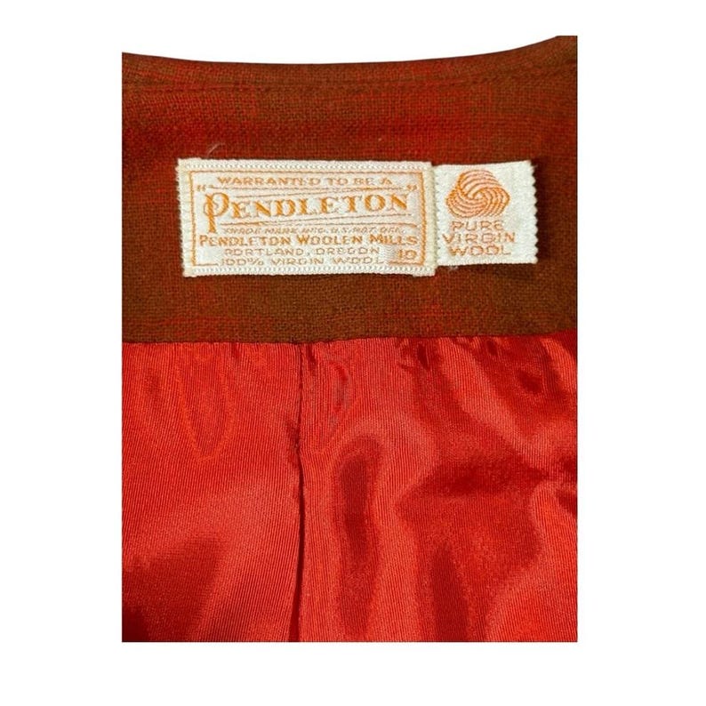 May include: Close-up of a brown garment interior with a bright red lining. A rectangular tag reads "WARRANTED TO BE A PENDLETON" and "PENDLETON WOOLEN MILLS PORTLAND, OREGON 100% VIRGIN WOOL". A smaller tag reads "PURE VIRGIN WOOL".