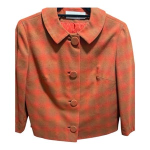 May include: A cropped, orange and brown plaid blazer with a rounded collar and three matching buttons. The blazer has a tailored fit and a solid orange lining. The jacket is hanging on a black hanger.
