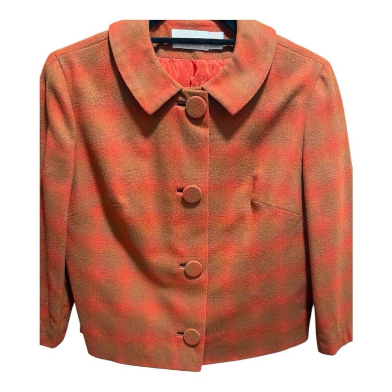 May include: A cropped, orange and brown plaid blazer with a rounded collar and three matching buttons. The blazer has a tailored fit and a solid orange lining. The jacket is hanging on a black hanger.