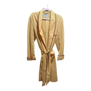 May include: A light yellow robe with brown trim and a matching belt. The robe is hanging on a pink hanger against a white background. The robe has long sleeves and a shawl collar.