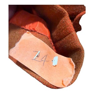 May include: Close-up of a section of a brown and orange garment. A light orange tag is attached with the handwritten number "24" and a small, light blue plastic tag. The garment has a textured appearance.