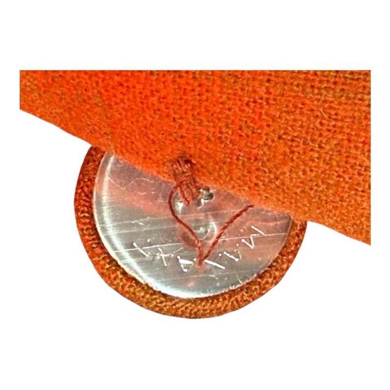 May include: Close-up of a clear, round button with the word "MA" etched into it, sewn onto a piece of orange fabric. The button is attached with brown thread, and the fabric has a textured appearance.