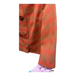 May include: A close-up of a vintage blazer in shades of orange and brown. The jacket features a textured pattern and three round buttons. The sleeve is visible, and the fabric appears to be wool or a similar material.