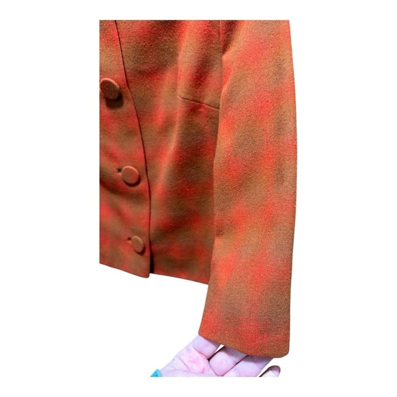 May include: A close-up of a vintage blazer in shades of orange and brown. The jacket features a textured pattern and three round buttons. The sleeve is visible, and the fabric appears to be wool or a similar material.