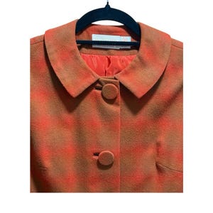 May include: A vintage orange and brown plaid jacket with a round collar and two matching buttons. The jacket is hanging on a black hanger. The lining is a solid orange color.