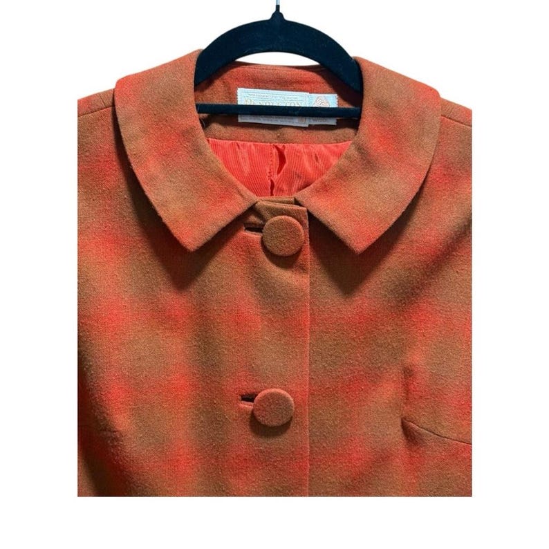 May include: A vintage orange and brown plaid jacket with a round collar and two matching buttons. The jacket is hanging on a black hanger. The lining is a solid orange color.