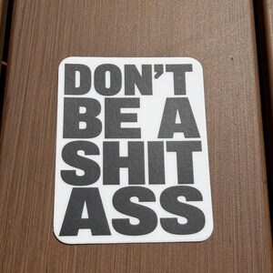 May include: A rectangular sticker with rounded corners featuring the text DON'T BE A SHIT ASS in bold, black, sans-serif capital letters centered on a white background. The sticker has a clean, flat design with high contrast between the black text and white.